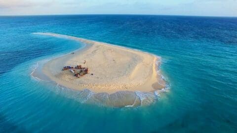 Aerial view of a small, sandy island surrounded by blue ocean waters with a group of people gathered in the center, enjoying an ecotourism tour.