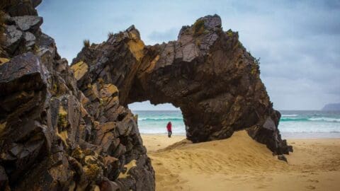 A person in a red coat stands on a sandy beach near a large rock formation with an arch, facing the sea under a cloudy sky, taking in the beauty of nature.