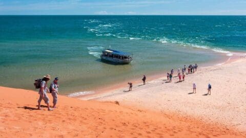 A group of people stand and walk on a sandy beach by azure waters, with a boat moored close to the shore. Two individuals are seen ascending the sandy dune in the foreground, perhaps exploring eco-friendly accommodations nearby.