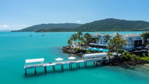 A coastal resort with a long pier extending over the turquoise waters of the Coral Sea, surrounded by palm trees under a clear blue sky. Hills are visible in the background, adding to the breathtaking view.