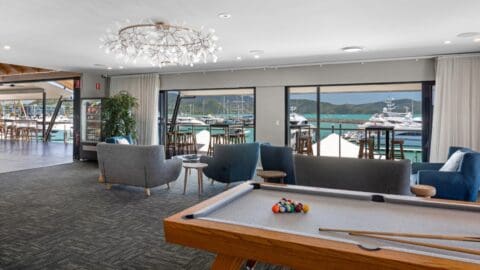 A lounge area with a pool table, comfortable seating, and large windows offering picturesque views of the Coral Sea marina adorned with elegant yachts.