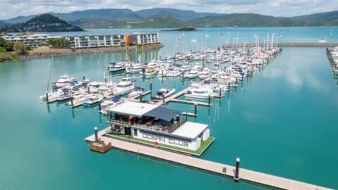 A charming marina with multiple docked boats and a floating building labeled "Ocean Club" rests on the turquoise waters of the Coral Sea. Hills and buildings grace the background under a partly cloudy sky, creating an inviting resort-like atmosphere.