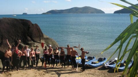 A group of people in wetsuits stands on a beach beside several kayaks, embracing an ecotourism adventure. The ocean and islands are visible in the background under a clear blue sky.