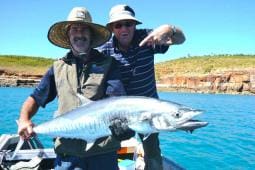 Two people on a charter boat holding a large fish, with a clear blue sky and water in the background. One person points at the fish. Both are wearing hats and sunglasses, enjoying their company out on the water.