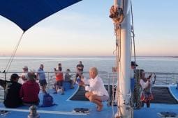 People are on the deck of a charter boat during sunset, some using their phones to take photos. The ocean and horizon are visible in the background, with occasional glimpses of sealife adding to the serene scene.
