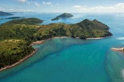 Aerial view of several lush green islands in the Whitsunday region, surrounded by clear blue ocean water under a partly cloudy sky. Perfect for a yacht rent adventure.