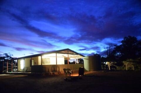 A single-story building with illuminated interior lights sits under a deep blue evening sky in the serene Bendleby Ranges, surrounded by a few trees and outdoor chairs. A large cylindrical water tank stands beside the building.