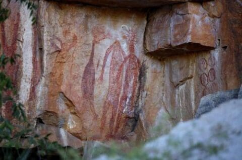 Ancient rock art with red ochre figures painted on a rocky surface, depicting human-like shapes and geometric patterns. The scene is located outdoors amidst rocky terrain, often explored during Nitmiluk Tours.