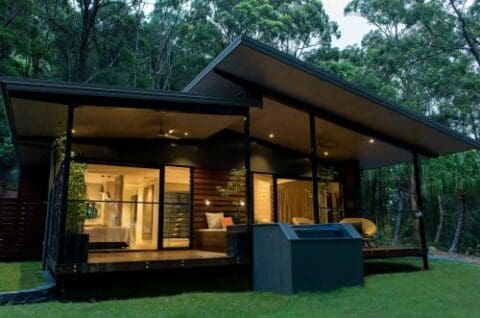 A modern house with large glass windows, a slanted roof, and a covered porch, set against a backdrop of dense trees. The interior is warmly lit, revealing a living area and bedroom that exudes the serene lifestyle of a Gwinganna retreat.