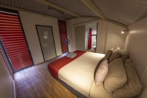 A small, neatly arranged bedroom at Savannah Lodge features a double bed, red and white bedding, a fan, red window blinds, a wooden floor, and an adjacent bathroom visible through an open doorway.