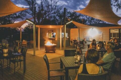 Outdoor restaurant with people dining around tables and a central fire pit at twilight, reminiscent of the rustic charm you'd find at Savannah Lodge, all covered by elegant shaded structures.