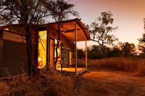 A small cabin with a corrugated metal roof, resembling the rustic charm of Savannah Lodge, is illuminated from within during sunset, set in a rural, grassy landscape with surrounding trees.