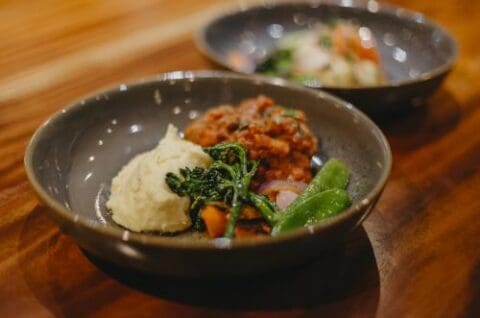 A dark gray bowl on a wooden table containing mashed potatoes, sautéed greens, and a red stew, reminiscent of a hearty meal enjoyed at a serene lodge near the Bungle Bungle range, with another similar dish blurred in the background.