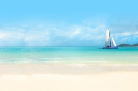 A white sailboat glides on clear turquoise waters near a sandy beach under a blue sky with scattered clouds, perfect for a Whitsundays cruise.