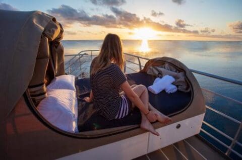 A person sits on a cushioned outdoor bed on a boat's deck, facing the ocean at sunset during a Whitsundays cruise. Towels are folded beside them.