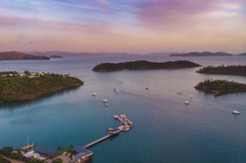 Aerial view of a coastal marina with yachts docked and islands surrounded by blue water under a cloudy sky, offering an ideal setting to rent a boat in the stunning Whitsunday region.
