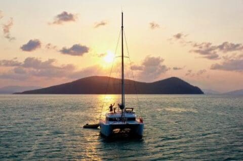 A yacht on calm waters at sunset, with two people standing on deck. Background includes a setting sun, partly cloudy sky, and hilly islands. Perfect for exploring the Whitsunday Islands.