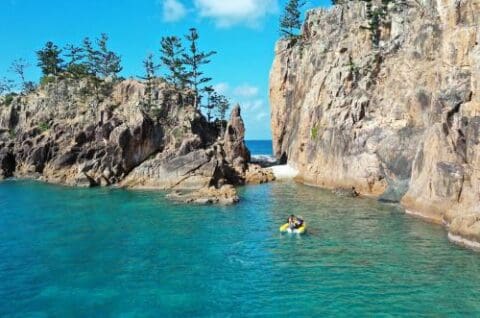 Two people kayaking in a clear, turquoise sea surrounded by rocky cliffs and pine trees under a blue sky, while a white yacht floats nearby. The serene atmosphere embodies the beauty of the Whitsundays, making it the perfect spot to rent a kayak for an unforgettable adventure.