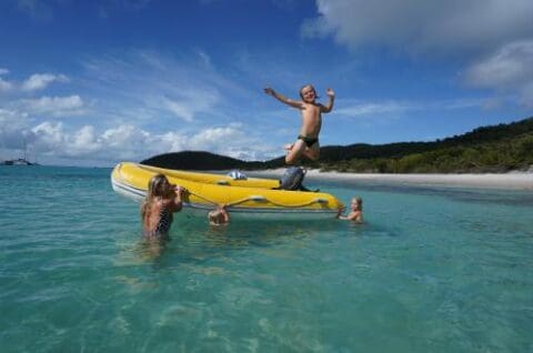 A child jumps off a yellow inflatable boat into clear, shallow water while an adult and two children watch nearby on a sunny day with a scenic Whitsunday beach and forested background.