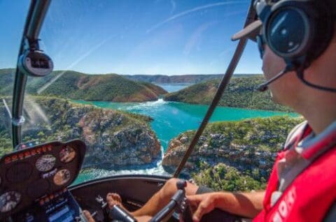 A person wearing a headset pilots a helicopter over the scenic landscape of Kimberley, with green hills, turquoise water, and a narrow waterway visible through the helicopter's windshield.