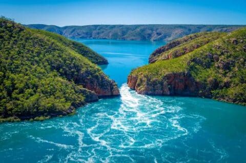Aerial view of Horizontal Falls in Australia, featuring turquoise water rushing through rocky cliffs surrounded by lush green hills under a clear blue sky—a spectacular sight often explored on Kimberley Cruises.