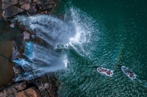 Aerial view of a waterfall cascading into a body of water, with two boats carrying people nearby. Rocks are visible near the waterfall. Experience this natural wonder on Kimberley Cruises aboard the Discovery One.