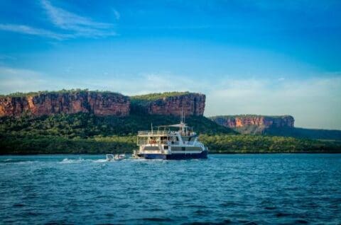 A boat from Kimberley Cruises sails on a body of water with two large cliffs and dense greenery in the background under a clear blue sky.