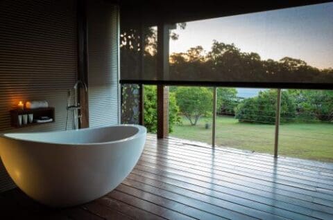 A white freestanding bathtub graces an outdoor wooden deck, offering a seamless retreat with views of a grassy yard and trees in the background—perfect for your tranquil lifestyle at Gwinganna.