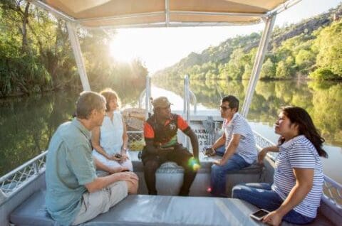 Five people sit and converse on a small boat on a calm river surrounded by lush greenery, with the sun setting in the background, reminiscent of serene Nitmiluk tours.