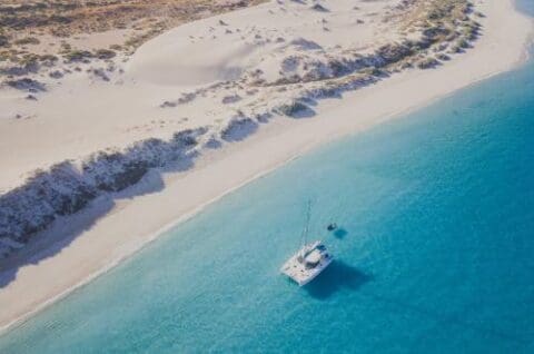 Aerial view of a Sail Ningaloo catamaran anchored in clear blue waters near a sandy beach with dunes and sparse vegetation.