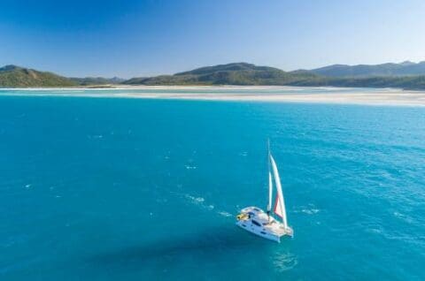 A rented yacht glides on clear, blue water with a sandy beach and mountains in the background under a Whitsunday sky.