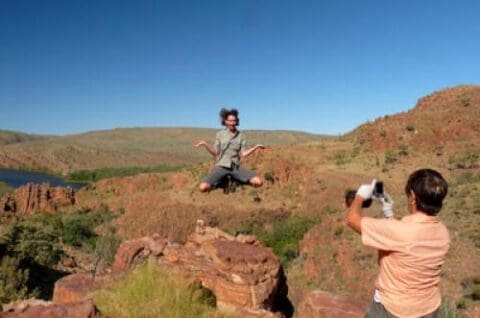 A person jumps mid-air on rocky terrain while another captures the moment; Willis Walkabouts in the background showcase a hilly landscape under a clear blue sky.
