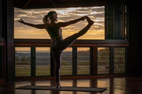 A woman practices yoga inside a room during sunset at Gwinganna, balancing on one leg with the other leg extended and holding her foot. Large windows in the background offer a view of trees and fields, creating an idyllic lifestyle retreat setting.