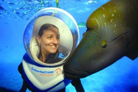 A certified person in an underwater helmet with "Sunlover Reef Cruises" branding interacts closely with a large fish in the ocean, showcasing the beauty of ecotourism.