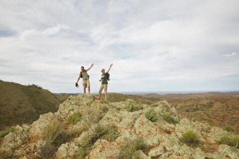 Two hikers stand on a rocky hilltop with arms raised, looking out over the vast, rugged landscape of the Bendleby Ranges under a cloudy sky.