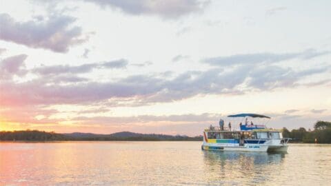 A tour boat sails on calm waters during sunset, with an open sky and a distant hilly landscape in the background, promoting ecotourism and sustainable travel.