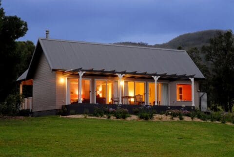 A single-story house with a sustainable metal roof and a lit-up veranda is surrounded by a grassy yard and trees, with hills in the background during dusk.