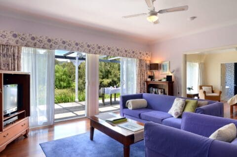 A living room with a blue couch, armchair, and a TV stand, featuring large glass doors leading to an outdoor deck with a view of greenery. The space is designed with sustainable decor elements and has a ceiling fan for comfort.