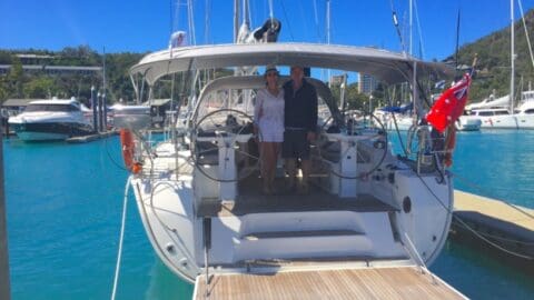 Two people stand on a docked sailboat in a vibrant marina, savoring their escape amidst clear blue waters and other boats gracefully anchored in the background.
