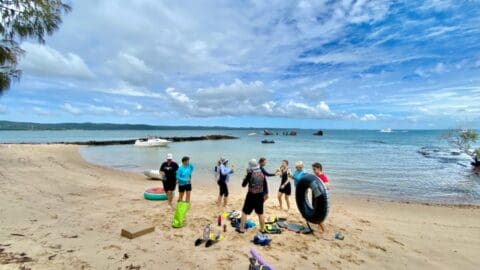 A group of people stands on a sandy beach, readying for Curlew Escape's water activities with gear like snorkeling equipment and a large rubber ring. Boats glide across the water in the background.