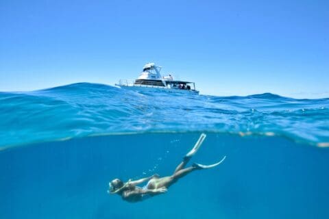 A woman wearing fins and a snorkel swims underwater near the surface, with a boat floating in the background under a clear blue sky. This scene highlights the beauty of ecotourism practices that emphasize harmony with nature and minimize environmental impact.