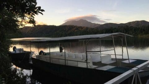 A touring boat, part of an ecotourism expedition, is docked at a lake surrounded by forested hills at dusk. Another smaller boat is seen in the background.