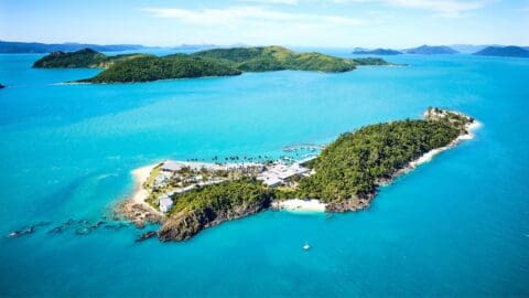 Aerial view of a tropical island with lush greenery, white sandy beaches, and turquoise waters. Several buildings, a dock, and a lone sailboat are visible. Distant islands appear in the background, making it an ideal spot for ecotourism tours.