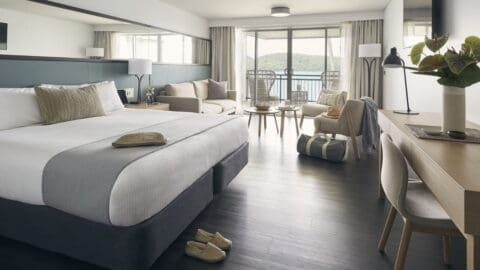 A modern hotel room with a large bed, armchairs, a desk, and a balcony offering views of the sea and hills. Decorated in neutral tones with a wooden floor, the room promotes sustainable tourism by featuring eco-friendly amenities throughout.