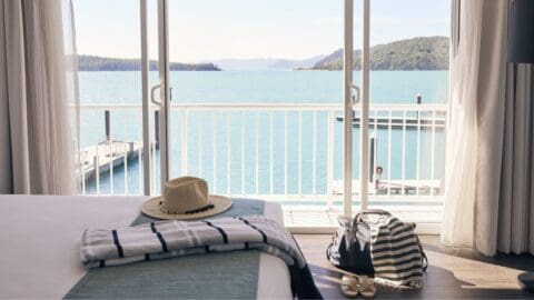 A serene waterfront view from a hotel room, featuring a bed with a striped blanket, a straw hat, and a striped bag with shoes on the floor. This ecotourism accommodation's balcony overlooks a dock and calm waters.