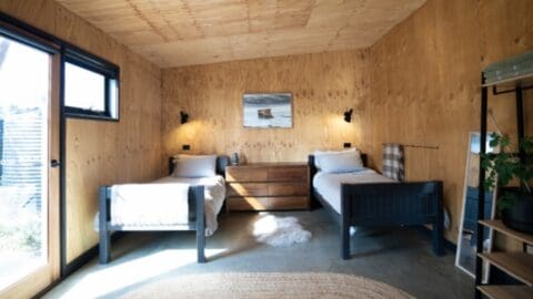 A small bedroom with two single beds, a wooden chest of drawers, and a framed picture on the plywood wall. A window and a glass door provide natural light and offer lovely views of nature.