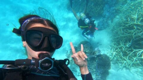 A young woman taking a selfie doing a rock sign while diving and another person taking pictures of coral on the sea ground.