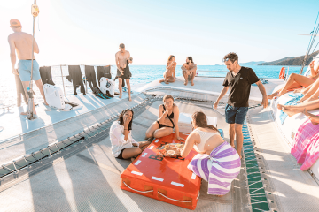 A group of people relax on a boat deck, some seated around a central red mat, while others stand or sit near the edge, enjoying a sunny day on the water during a guided tour.