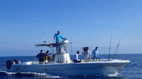 A group of people on a boat with "East Coast Angling" printed on the side are fishing in the ocean. Several fishing rods are set up, and one person is filming, capturing the essence of a guided tour in nature.