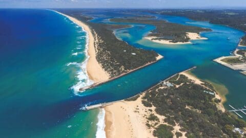 Aerial view of a coastal area featuring a long sandy beach, clear blue waters, an inlet with piers, and a backdrop of green vegetation and contrasting waterways.